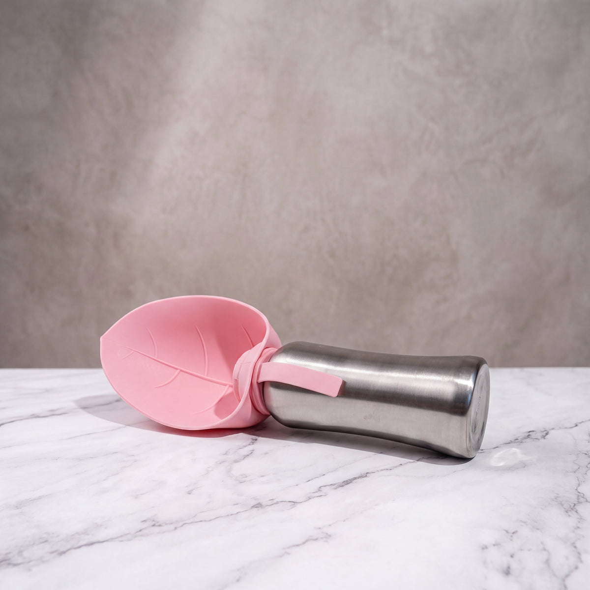 Gourde de voyage isotherme pour chien rose pastel allongée, écuelle en forme de feuille déployée sur une table en marbre.