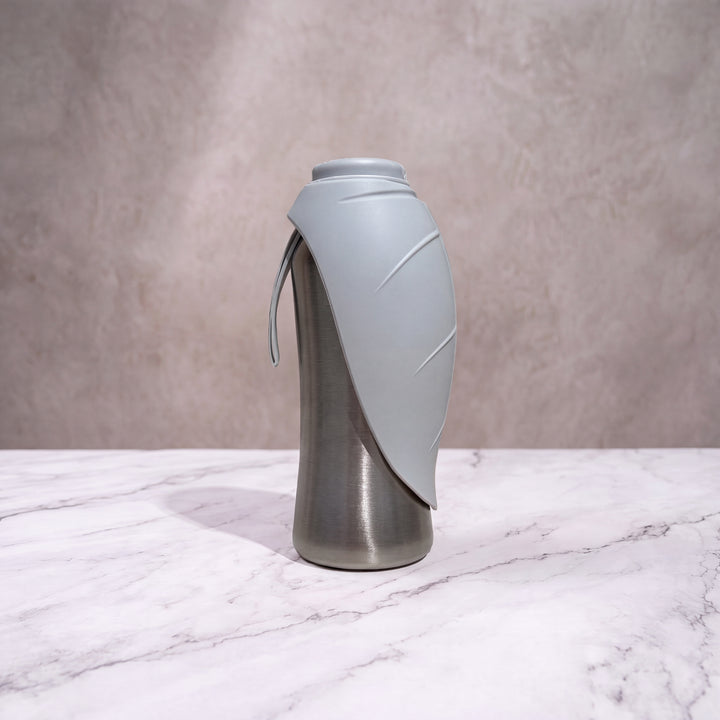 Gourde isotherme pour chien grise et inox de 750ml debout avec son écuelle feuille repliée sur fond marbré.